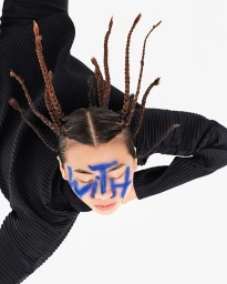 Portrait of a person with blue spray paint on face and braided hair in black ribbed top against white background