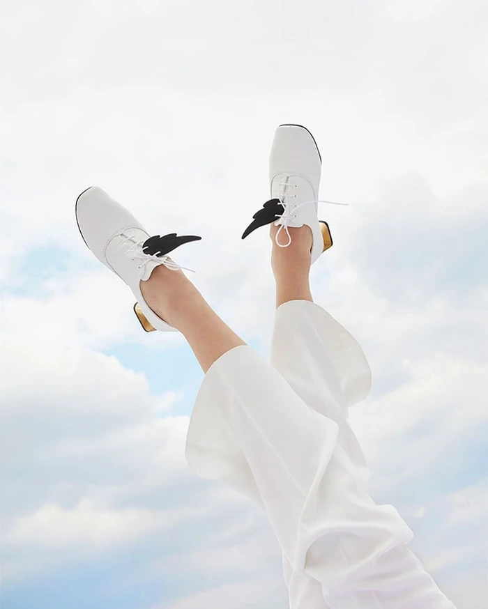 Legs elevated against a cloudy sky wearing white lace-up heeled shoes with black wing details and white trousers