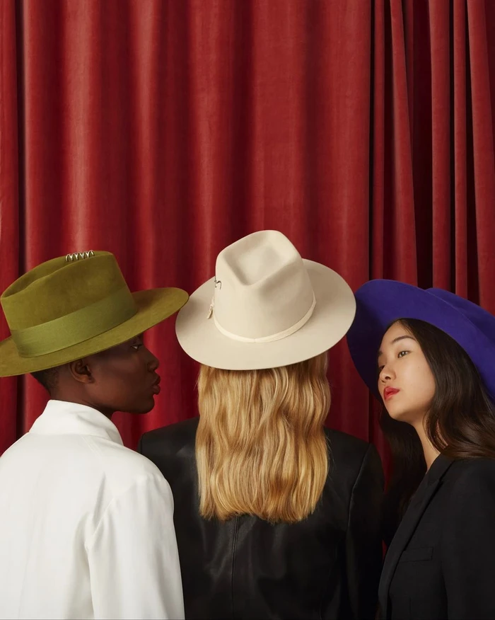 Three models wearing colorful fedoras and hats against a draped red curtain background, photographed by Laura Bonnefous.