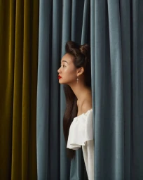 Profile of a woman with red lipstick parting blue and gold velvet curtains in a portrait by Laura Bonnefous
