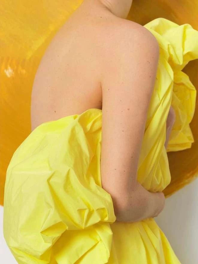 Close-up portrait of a person holding bright yellow crumpled fabric against a warm yellow and white background.