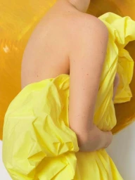 Close-up portrait of a person holding bright yellow crumpled fabric against a warm yellow and white background.