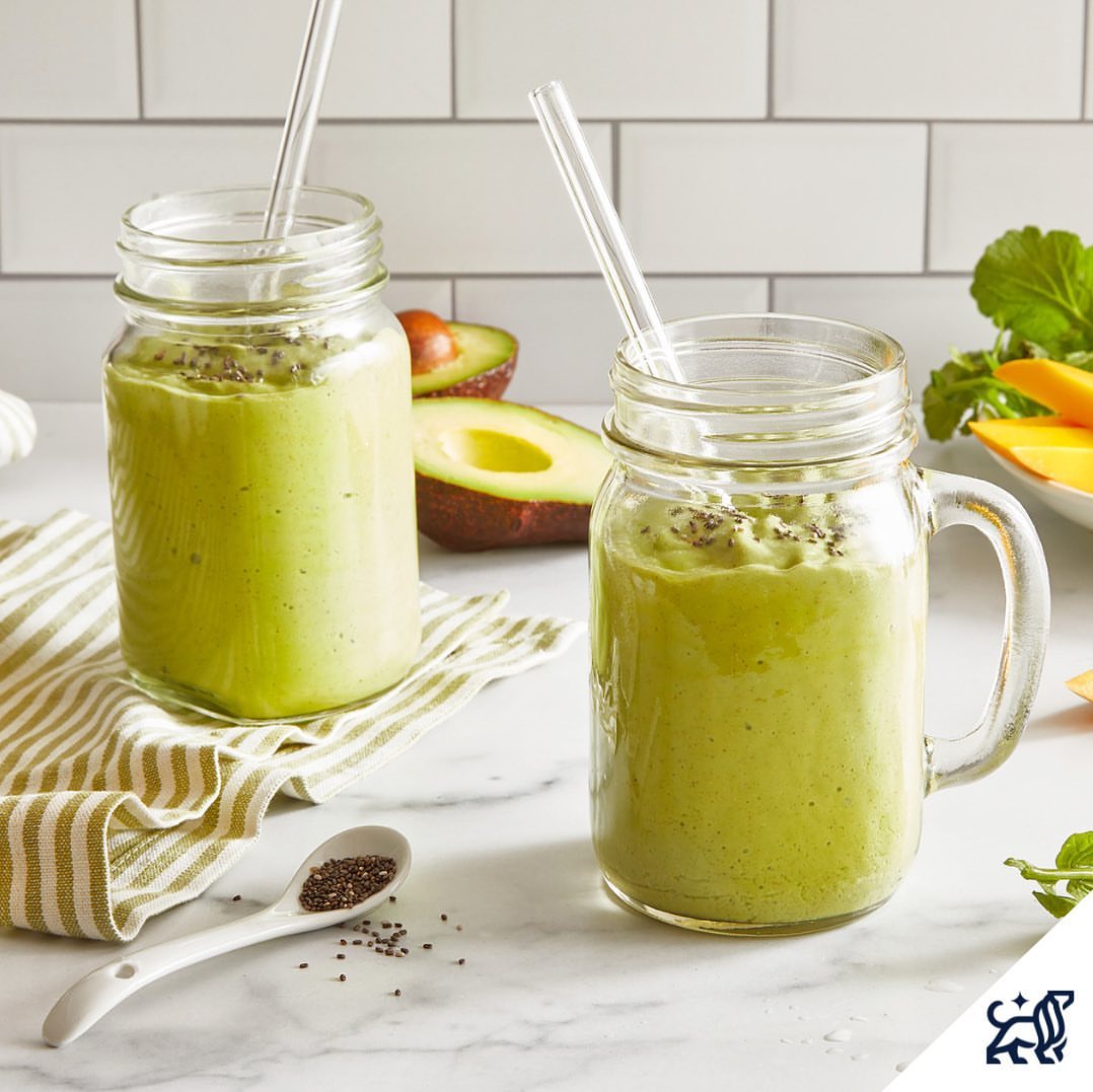 Two mason jars filled with vibrant green smoothies, each with a straw, sitting on a white marble countertop with avocados and greenery.
