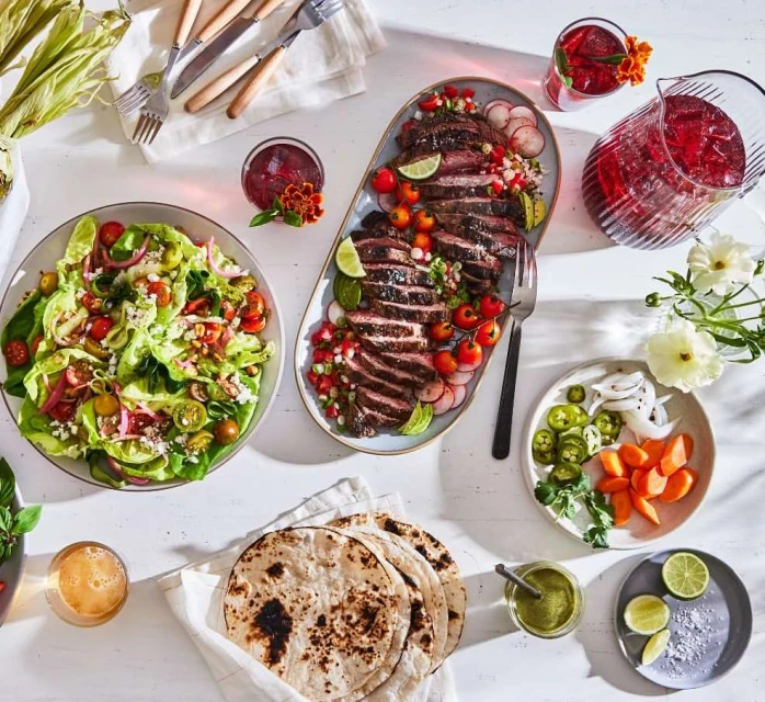 Overhead shot of a table spread with steak, salad, tortillas, and drinks, bathed in bright light, perfect for food and beverage advertising.