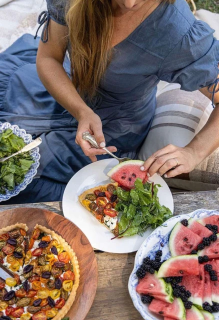 Woman in blue dress prepares watermelon and tomato tart for outdoor picnic with salad and blackberries on a wooden table.