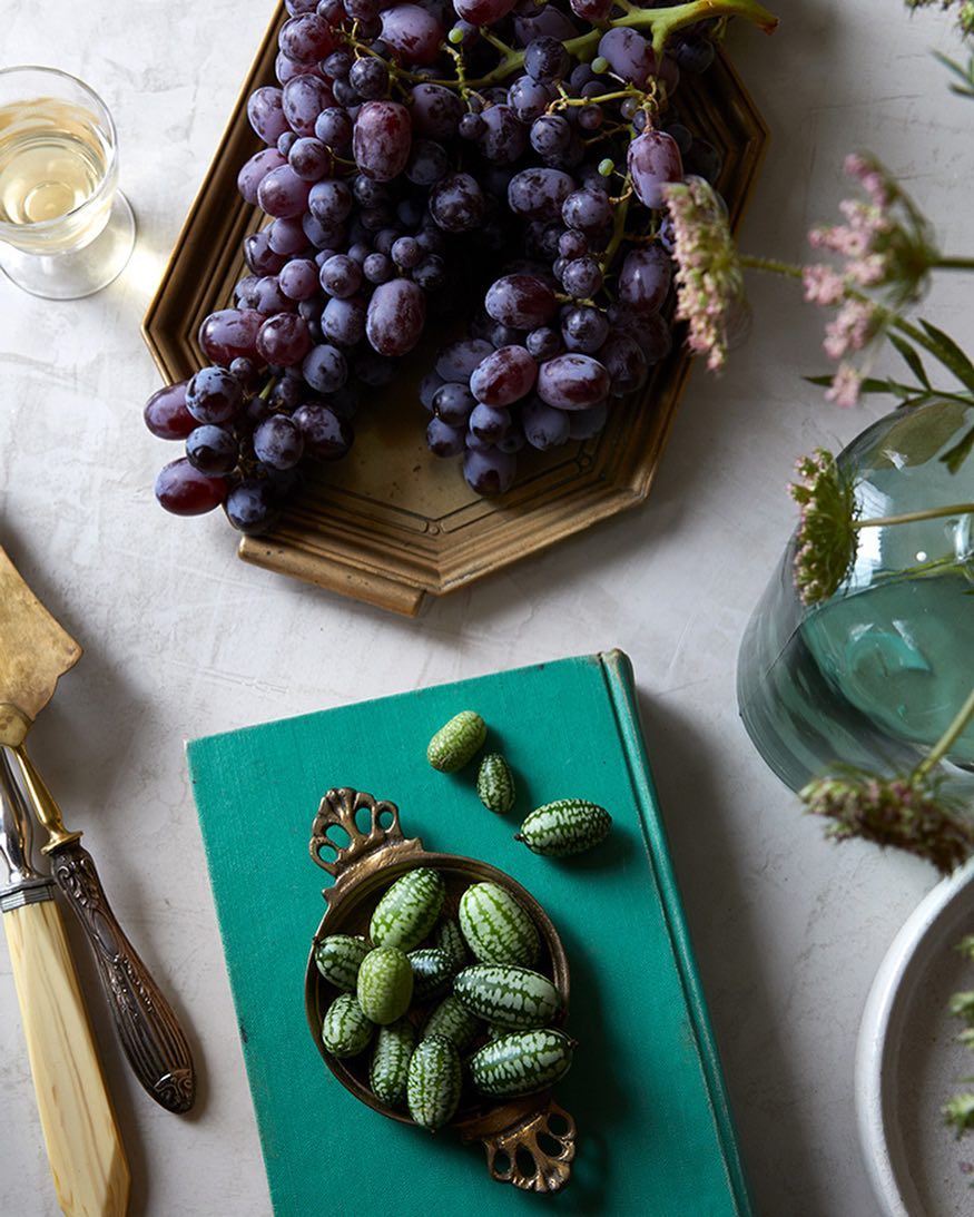 Overhead still life photo of dark grapes on a gold tray, green cucumbers in a bronze dish, and a glass of white wine on a grey surface.