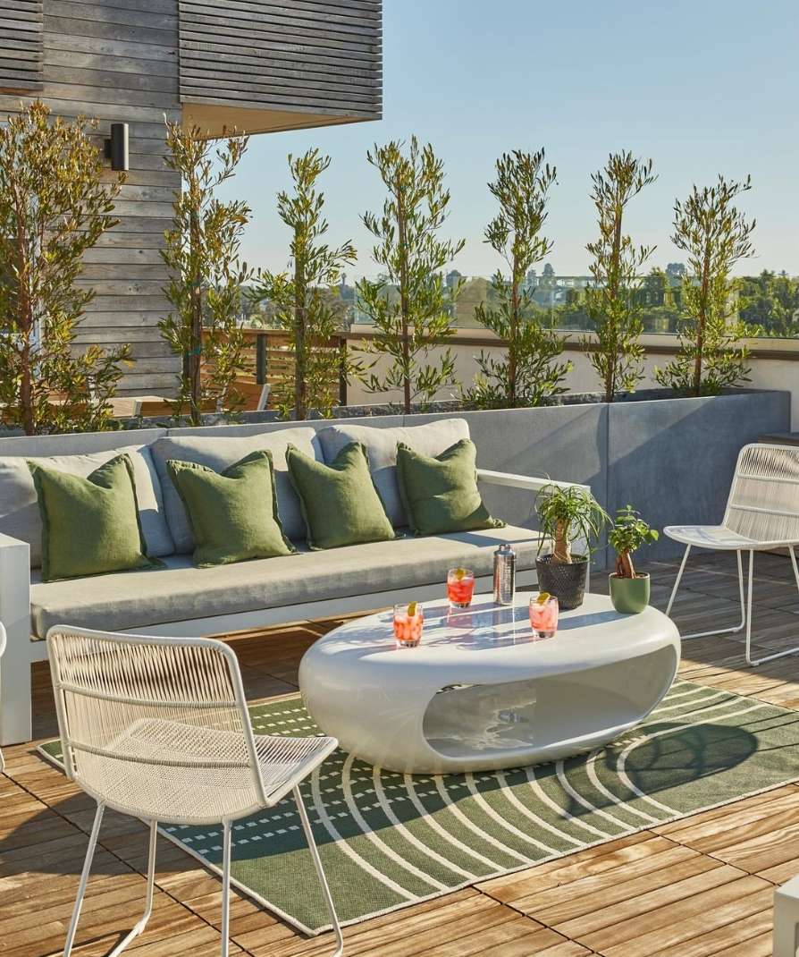 Outdoor patio scene featuring a gray sofa with green pillows, a white coffee table with drinks, and white chairs on a wooden deck.