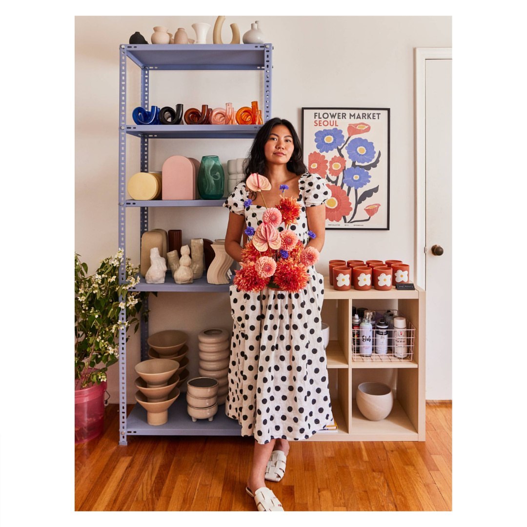 Young woman in polka-dot dress poses with a bouquet in front of a shelf of ceramic vases and bowls inside a home interior.