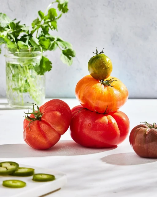 Tower of colorful heirloom tomatoes with fresh herbs in a glass jar, and sliced jalapenos on a cutting board. Studio lighting, food photography.