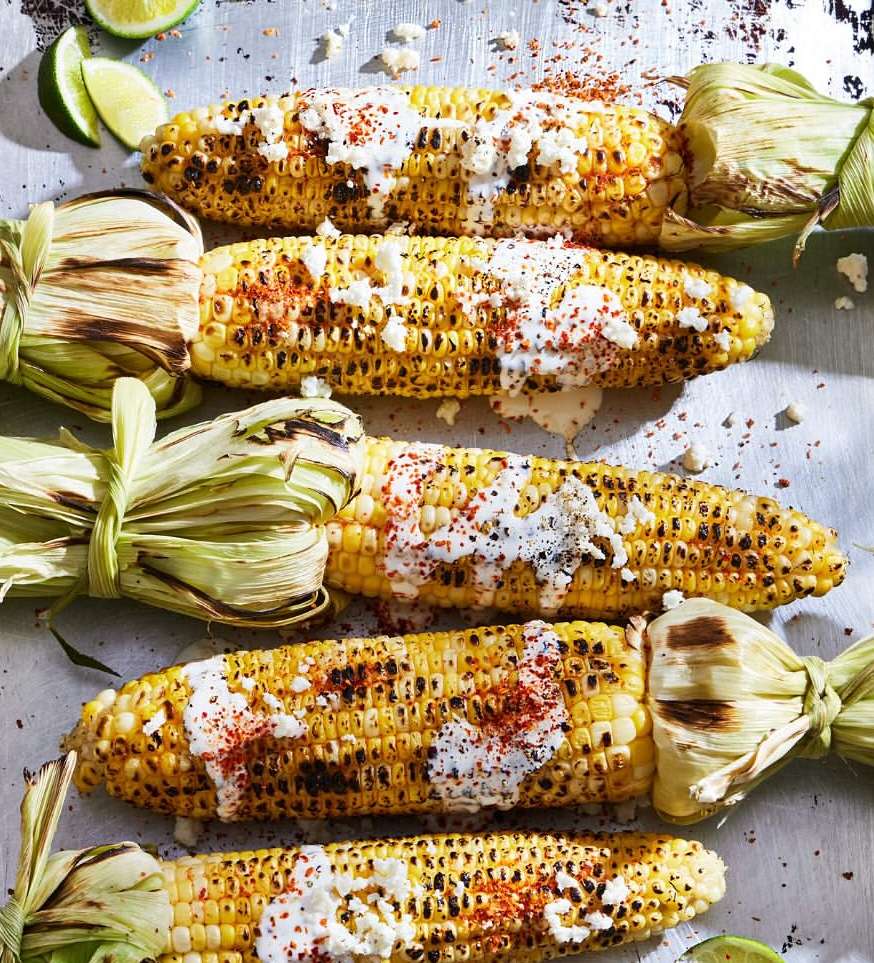 Grilled corn on the cob, Mexican street corn style, topped with crumbled cheese, chili powder, and lime wedges, arranged on a surface.