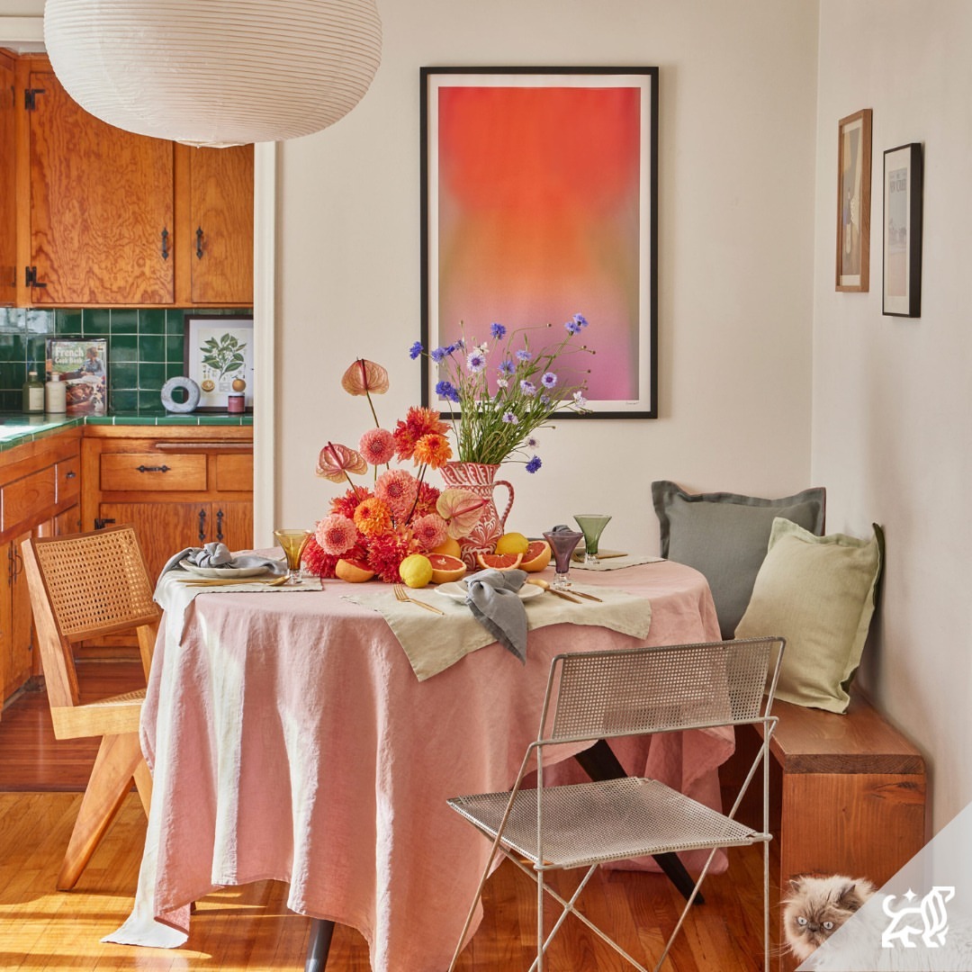 Interior home setting featuring a round dining table set with flowers and fruit, surrounded by chairs, with a cat peeking out from a bench.