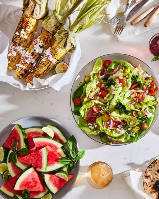Overhead shot of corn on the cob, salad, watermelon, lime, and drinks arranged on a white table, highlighting a fresh and colorful food presentation.
