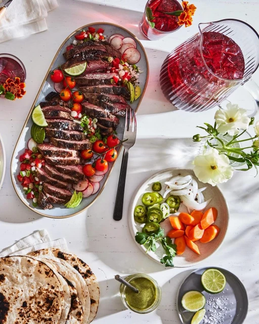 Overhead shot of sliced steak on a platter with tomatoes and radishes, accompanied by tortillas, salsa, and drinks on a white table.