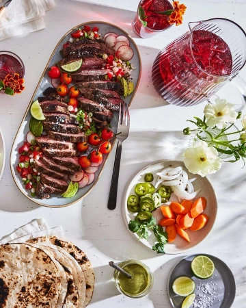 Overhead shot of sliced steak on a platter with tomatoes and radishes, accompanied by tortillas, salsa, and drinks on a white table.