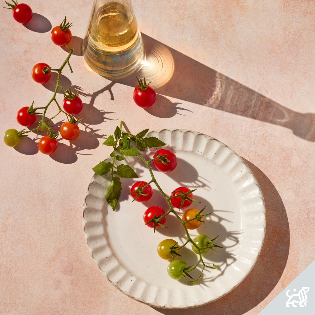 Overhead shot of cherry tomatoes on the vine arranged on a white plate with a bottle casting a shadow on a pink textured surface.