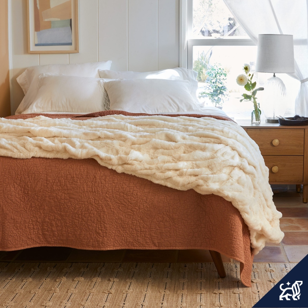 Bedroom features a bed with a rust-colored quilt, a cream throw blanket, and white pillows in a brightly lit interior setting.