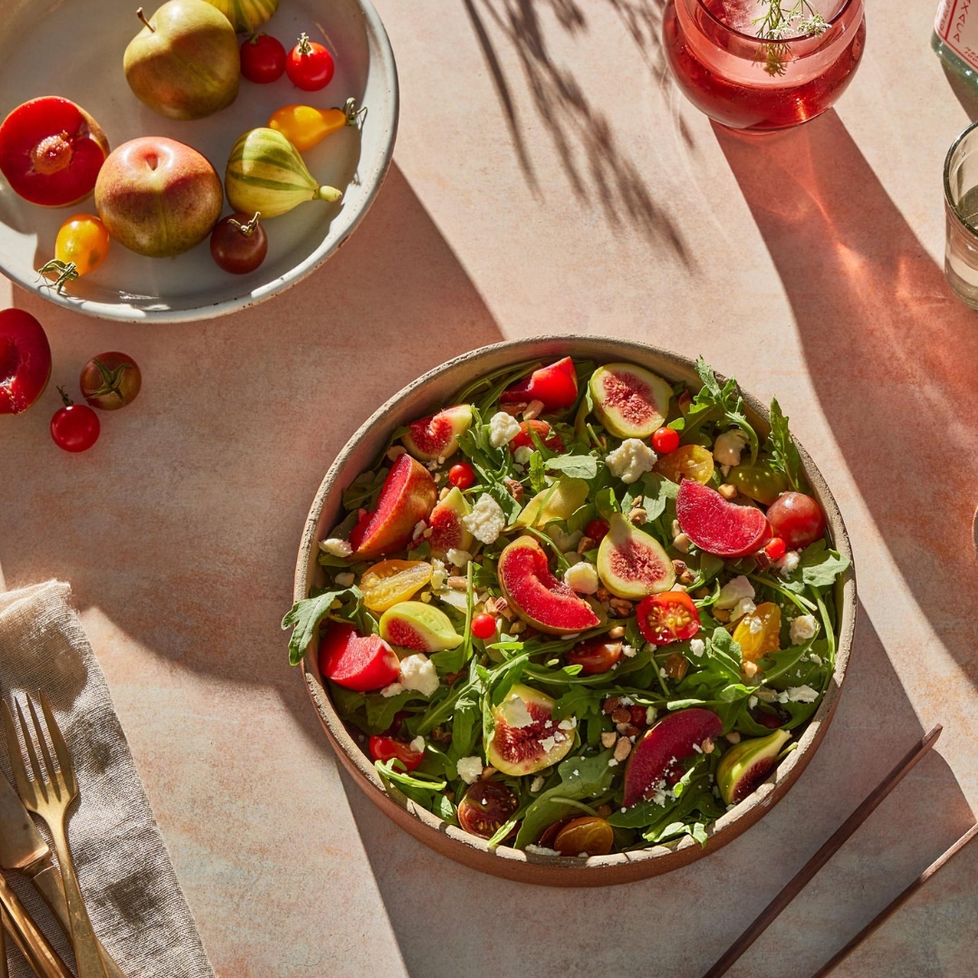 Overhead shot of a salad with figs, greens, tomatoes, and white cheese, plus a side of fruit on a pink surface with drinks.