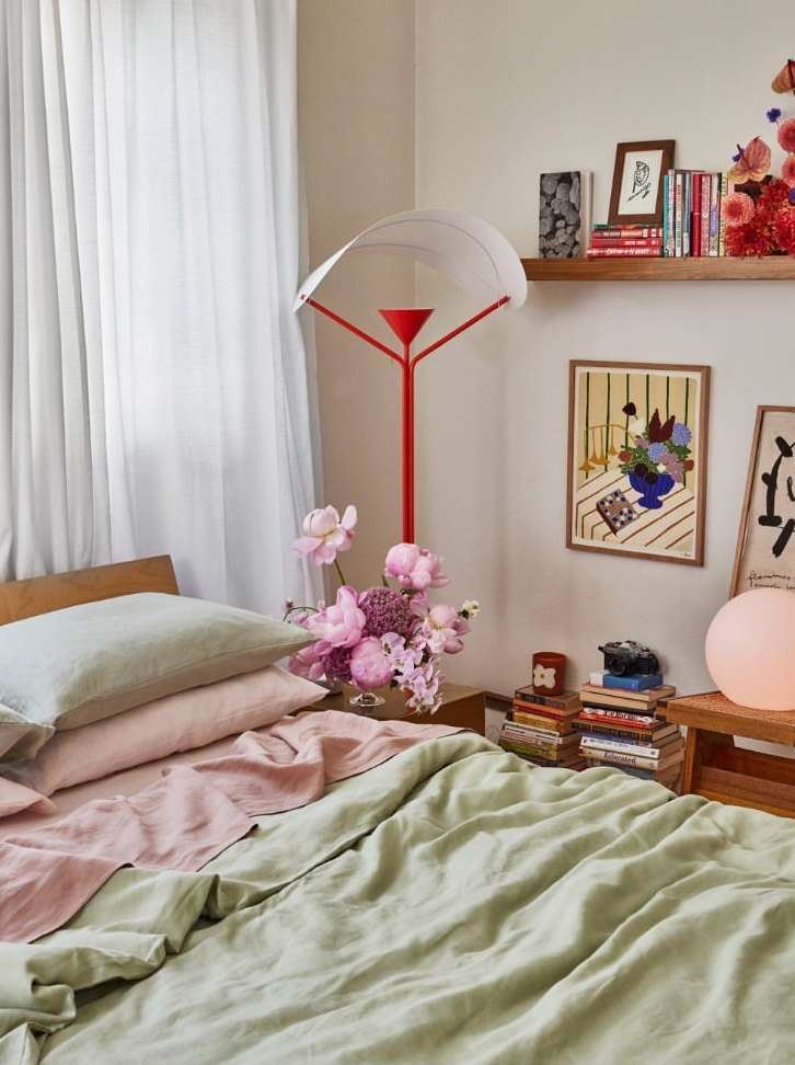 Bedroom interior with a bed covered in green and pink linens, a pink floral arrangement, books, art prints, and a floor lamp.