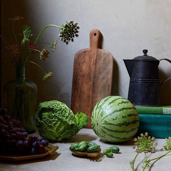 Still life composition featuring a watermelon, savoy cabbage, antique coffee pot, wooden cutting board, grapes, flowers, and books.