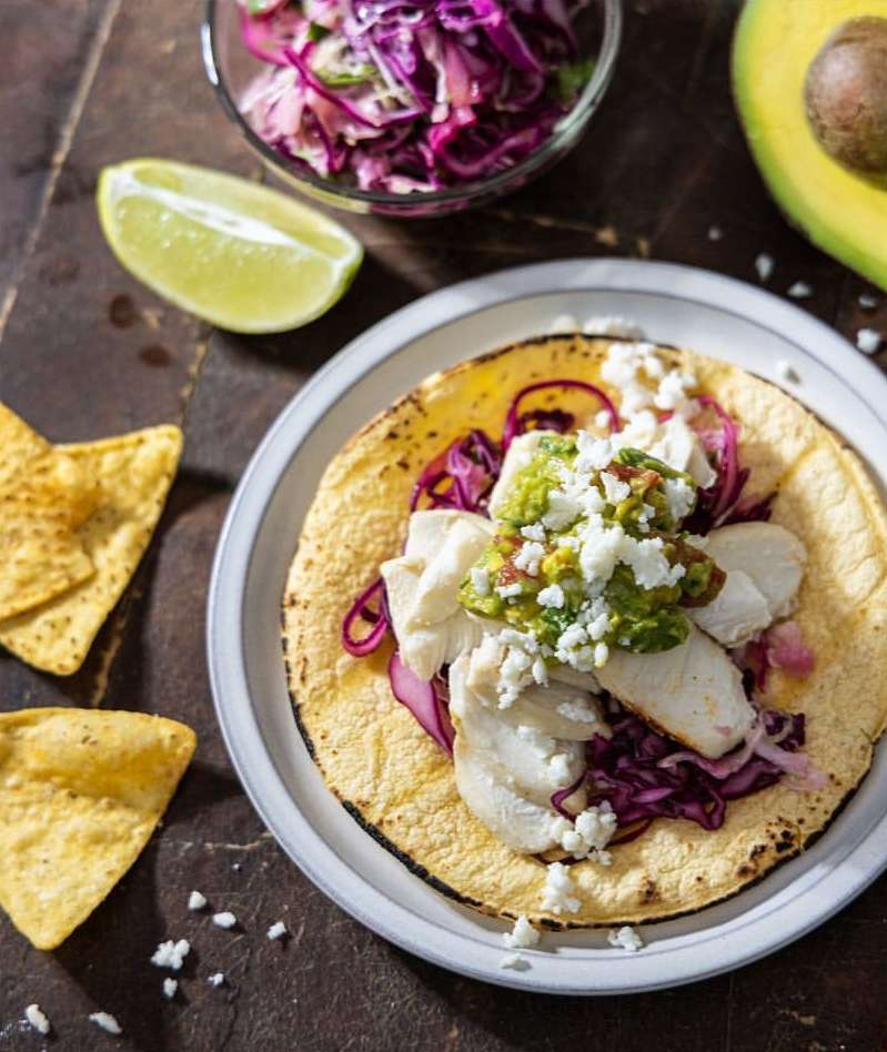 Overhead shot of a fish taco on a white plate with a lime wedge, chips, avocado, and cabbage slaw on a dark wooden surface.