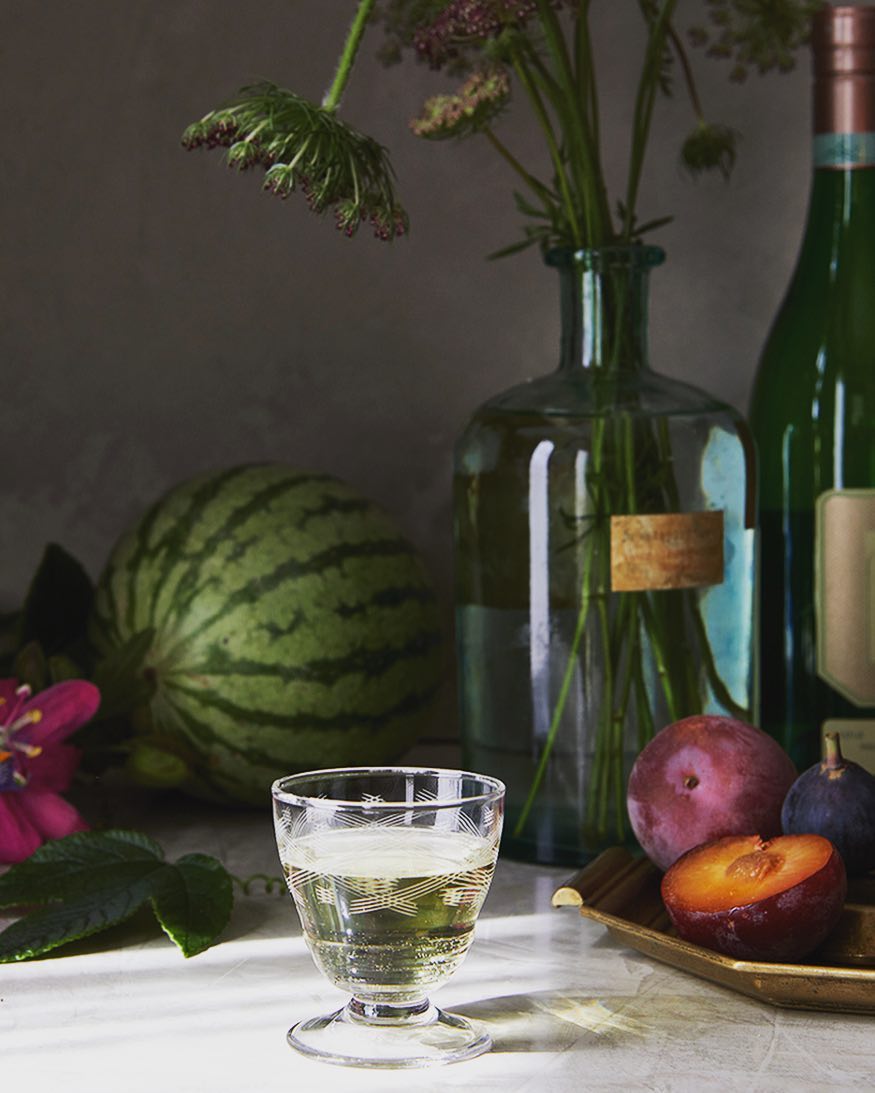 Still life shot of a glass of clear liquid, fruit, and green bottles against a muted grey background, with flowers in a glass vase.