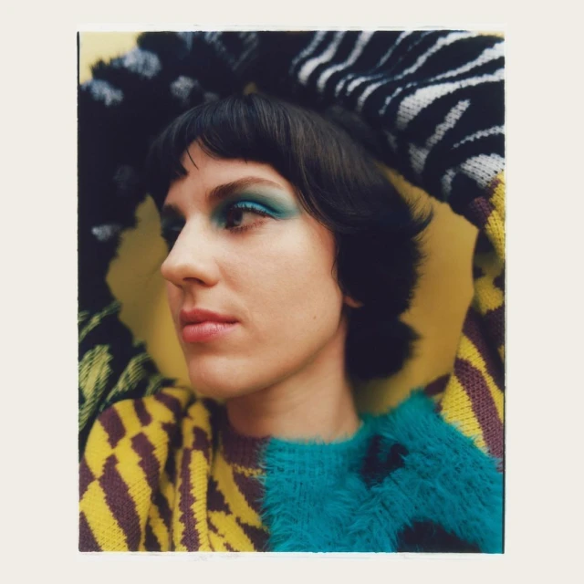 Portrait of woman with blue eye shadow wearing a patterned sweater against a yellow background in a studio setting. Fashion shot.