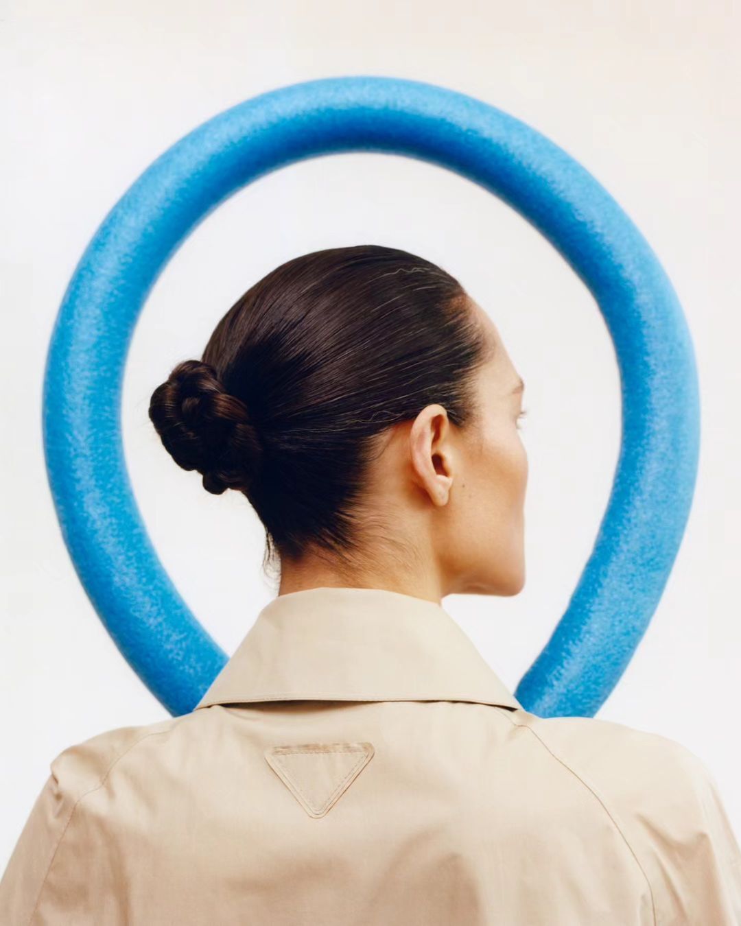 Woman in beige jacket with blue foam tube encircling her head. The jacket has a subtle triangle logo. Back view with white background.