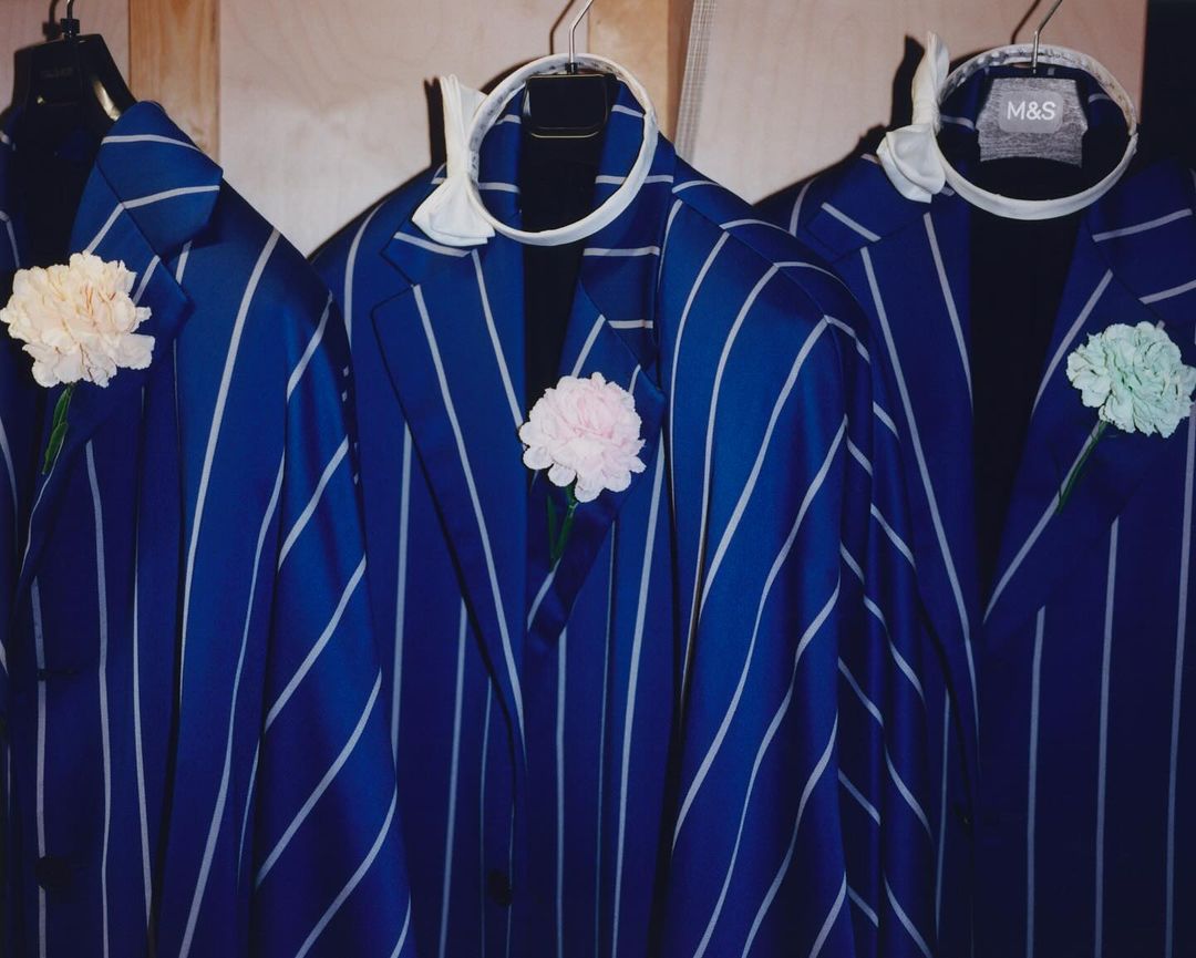 Three vibrant blue pinstripe suits with flower boutonnieres hang side-by-side on hangers, conveying formal fashion and tailoring.