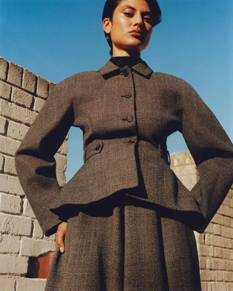 Woman in a gray tweed peplum jacket and skirt stands against a brick wall with blue sky above for a fashion editorial shot.