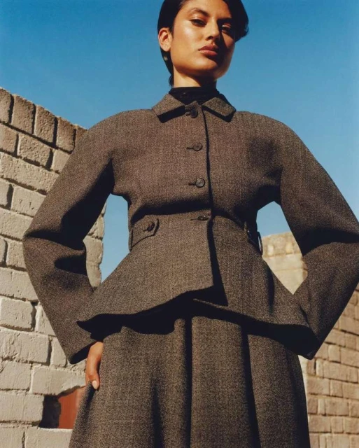 Woman in a gray tweed peplum jacket and skirt stands against a brick wall with blue sky above for a fashion editorial shot.