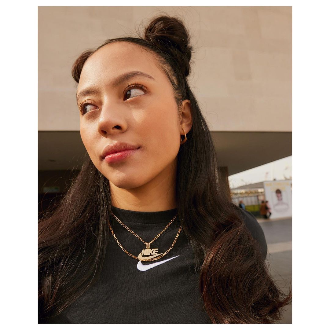 Portrait of a young woman with dark hair in space buns wearing a Nike T-shirt and gold necklaces, with a neutral facial expression.