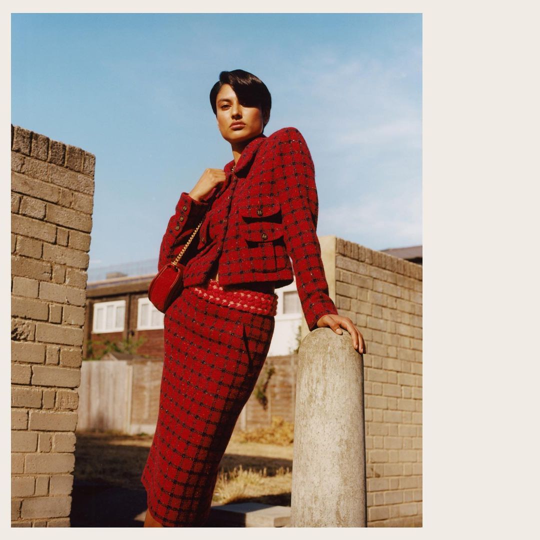 Fashion shot of an adult female in a red checkered skirt suit with a purse standing near a concrete wall in an urban outdoor environment.