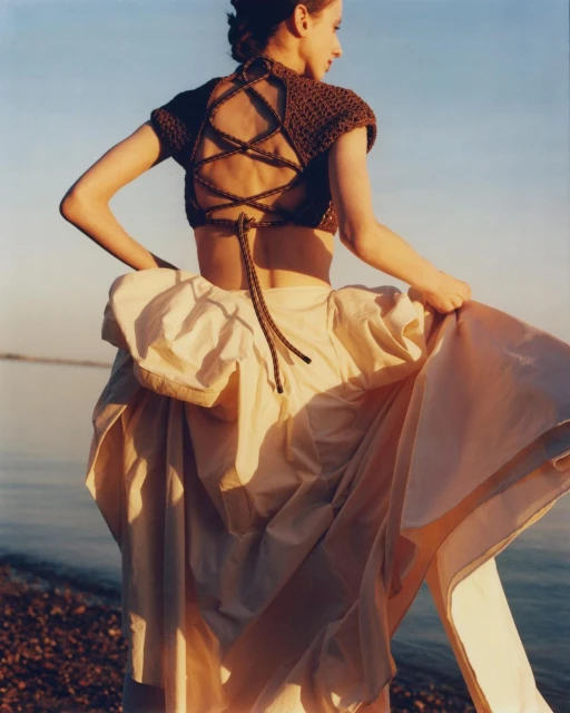 Woman in open-back brown crop top and tan flowing skirt stands on a beach, holding her skirt, facing away from the camera.