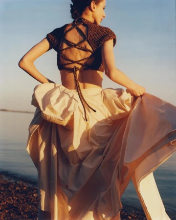 Woman in open-back brown crop top and tan flowing skirt stands on a beach, holding her skirt, facing away from the camera.