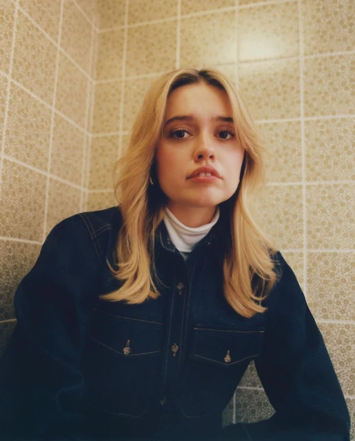Blonde woman in blue denim shirt and white turtleneck sweater sitting indoors with beige tiled wall in the background, looking at camera.