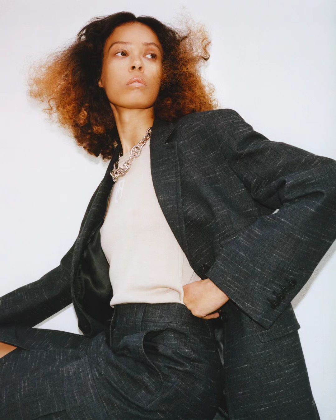 A woman with curly dark and blonde hair posing in a dark grey blazer and matching trousers in a studio setting with white background.