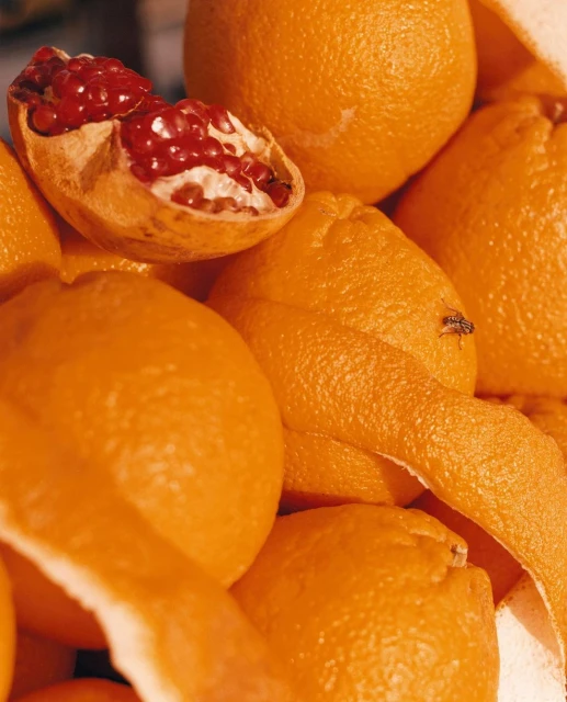 Close-up of a pile of vibrant oranges, one cut open revealing juicy red pomegranate seeds, creating a colourful citrus arrangement.