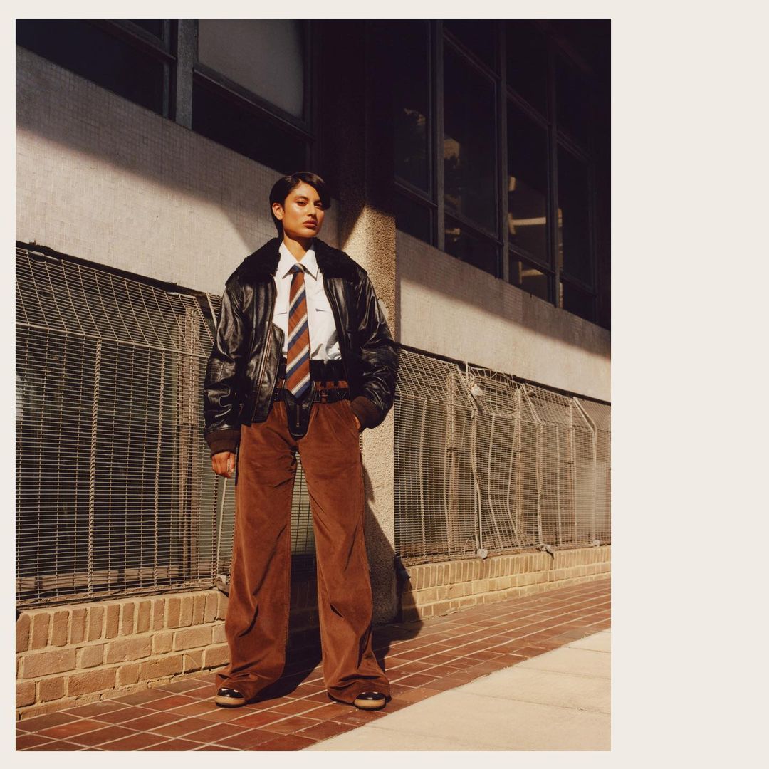An adult woman stands outside wearing a black jacket, white shirt, striped tie, brown trousers, and shoes against an urban background.