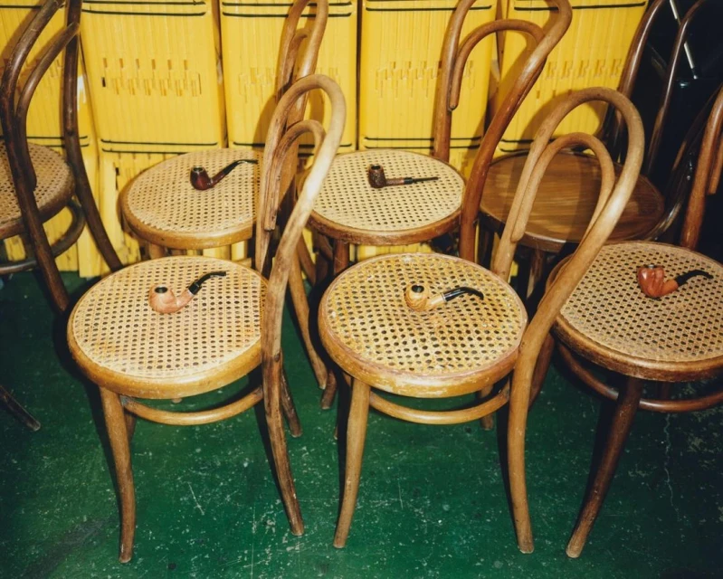 Several bentwood chairs with woven seats are arranged indoors, each holding a smoking pipe, against a yellow woven background.