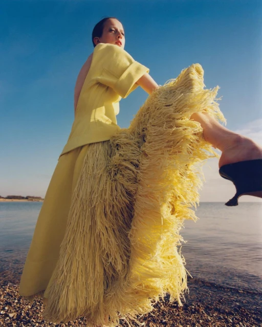 A woman in a yellow dress stands on a rocky beach with the ocean and blue sky in the background, creating a bright, summery fashion scene.