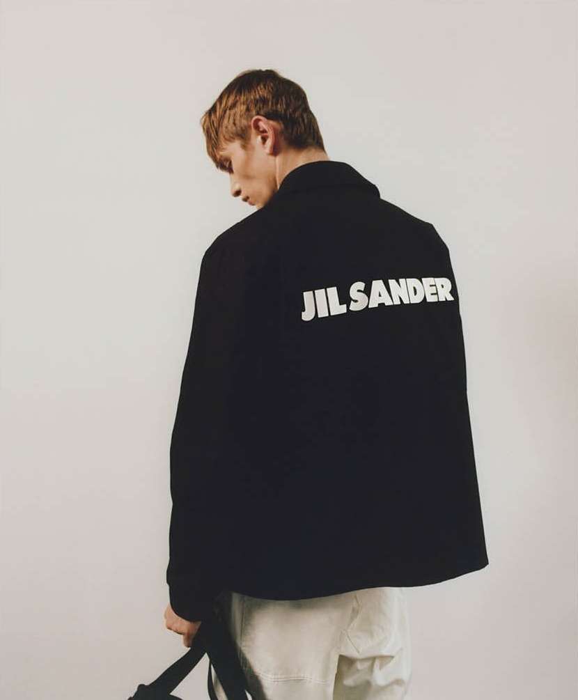A man wearing a black jacket with white Jil Sander lettering on the back against a neutral background, photographed by Laura Mccluskey.