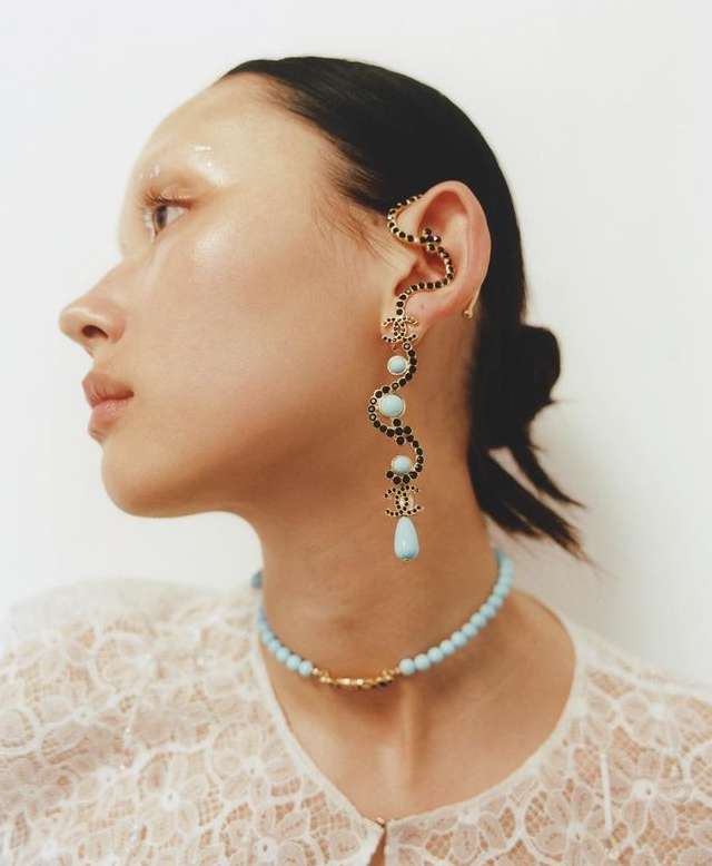 Profile of a woman with striking makeup and jewelry, including an ear cuff and beaded necklace, against a neutral backdrop.