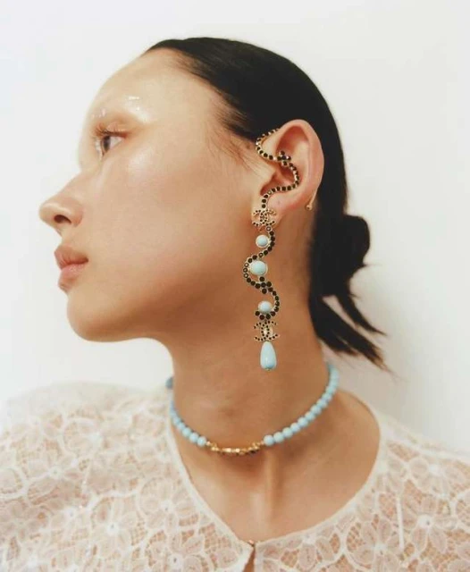 Profile of a woman with striking makeup and jewelry, including an ear cuff and beaded necklace, against a neutral backdrop.