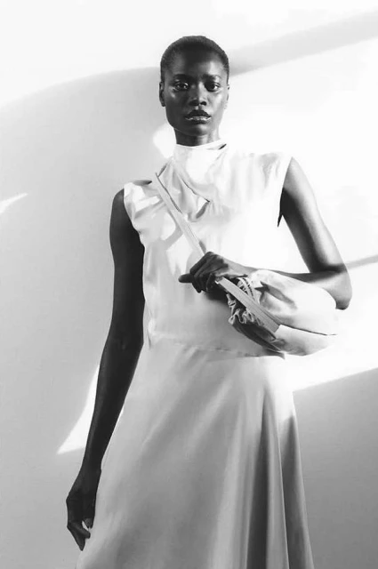 Monochrome portrait of a slender Black woman in a dress and carrying a handbag, posing against a light gray wall with sunlight.