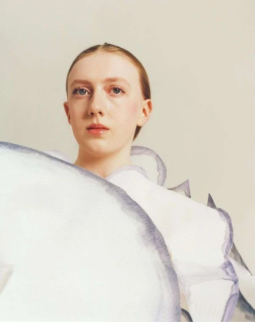 Portrait of a young woman in a white, translucent dress with lavender edges against a plain background in a photo studio.