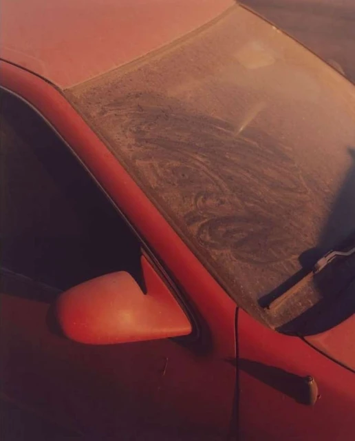 Close-up of a red car covered in dust with visible side mirror and windshield with dirt patterns; natural lighting. 