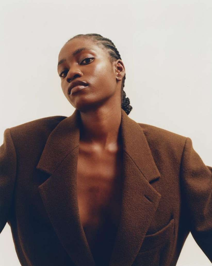 Elegant portrait of an African-American woman wearing a brown blazer against a plain white background in a studio fashion shot.
