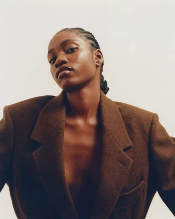 Elegant portrait of an African-American woman wearing a brown blazer against a plain white background in a studio fashion shot.
