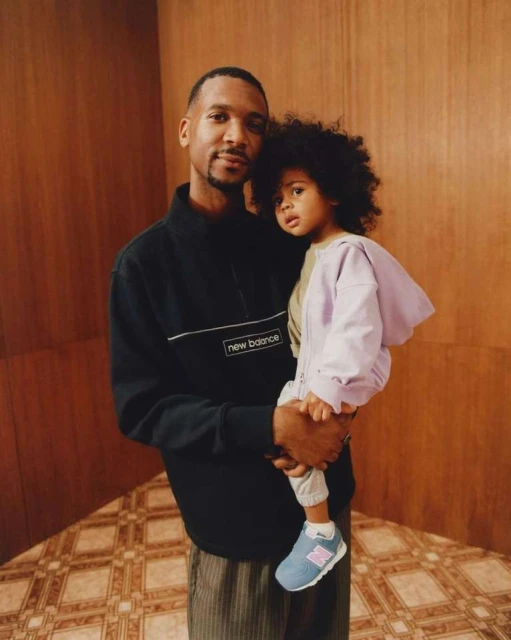 Portrait of a young African-American father holding his child, both wearing casual clothing, in a photo studio with wood-paneled backdrop.