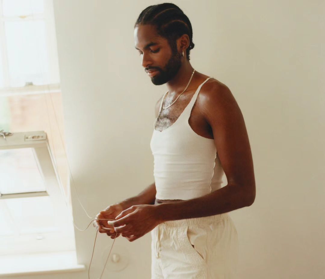 Portrait of an African American man in a white tank top and shorts standing indoors, holding wires, with natural light.